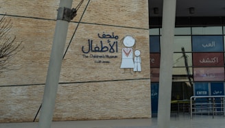A wall with a large sign for The Children's Museum in Arabic and English, featuring a simple drawing of a child and an adult in traditional clothing. The background is made of brick-like material, and the entrance with glass panels is visible.