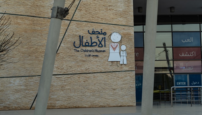 A wall with a large sign for The Children's Museum in Arabic and English, featuring a simple drawing of a child and an adult in traditional clothing. The background is made of brick-like material, and the entrance with glass panels is visible.