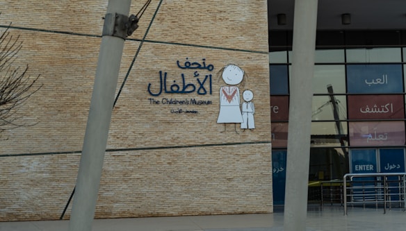 A wall with a large sign for The Children's Museum in Arabic and English, featuring a simple drawing of a child and an adult in traditional clothing. The background is made of brick-like material, and the entrance with glass panels is visible.