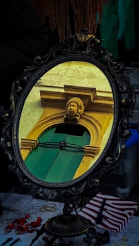 A decorative mirror with an ornate frame reflects an architectural element, featuring a carved head above a green wooden door with yellow stone surroundings. The reflection captures classical architectural details. Below the mirror, there is a table with various small objects, including some in red and white, possibly indicating a setting with personal or artistic items.