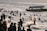 A large group of people are enjoying the water near the shore. Many are swimming or playing in the waves, while others are standing or walking along the beach. A boat is visible in the distance with a visible phone number and the name 'SISO' on it. The atmosphere is lively and crowded, indicative of a popular recreational area.