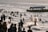 A large group of people are enjoying the water near the shore. Many are swimming or playing in the waves, while others are standing or walking along the beach. A boat is visible in the distance with a visible phone number and the name 'SISO' on it. The atmosphere is lively and crowded, indicative of a popular recreational area.