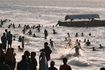 A large group of people are enjoying the water near the shore. Many are swimming or playing in the waves, while others are standing or walking along the beach. A boat is visible in the distance with a visible phone number and the name 'SISO' on it. The atmosphere is lively and crowded, indicative of a popular recreational area.