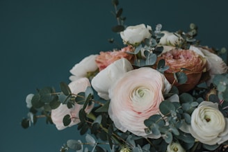 A bouquet of delicate flowers with pale pink and white petals surrounded by eucalyptus leaves. The soft colors of the flowers contrast with the dark green leaves, set against a deep green background, creating a serene and elegant composition.