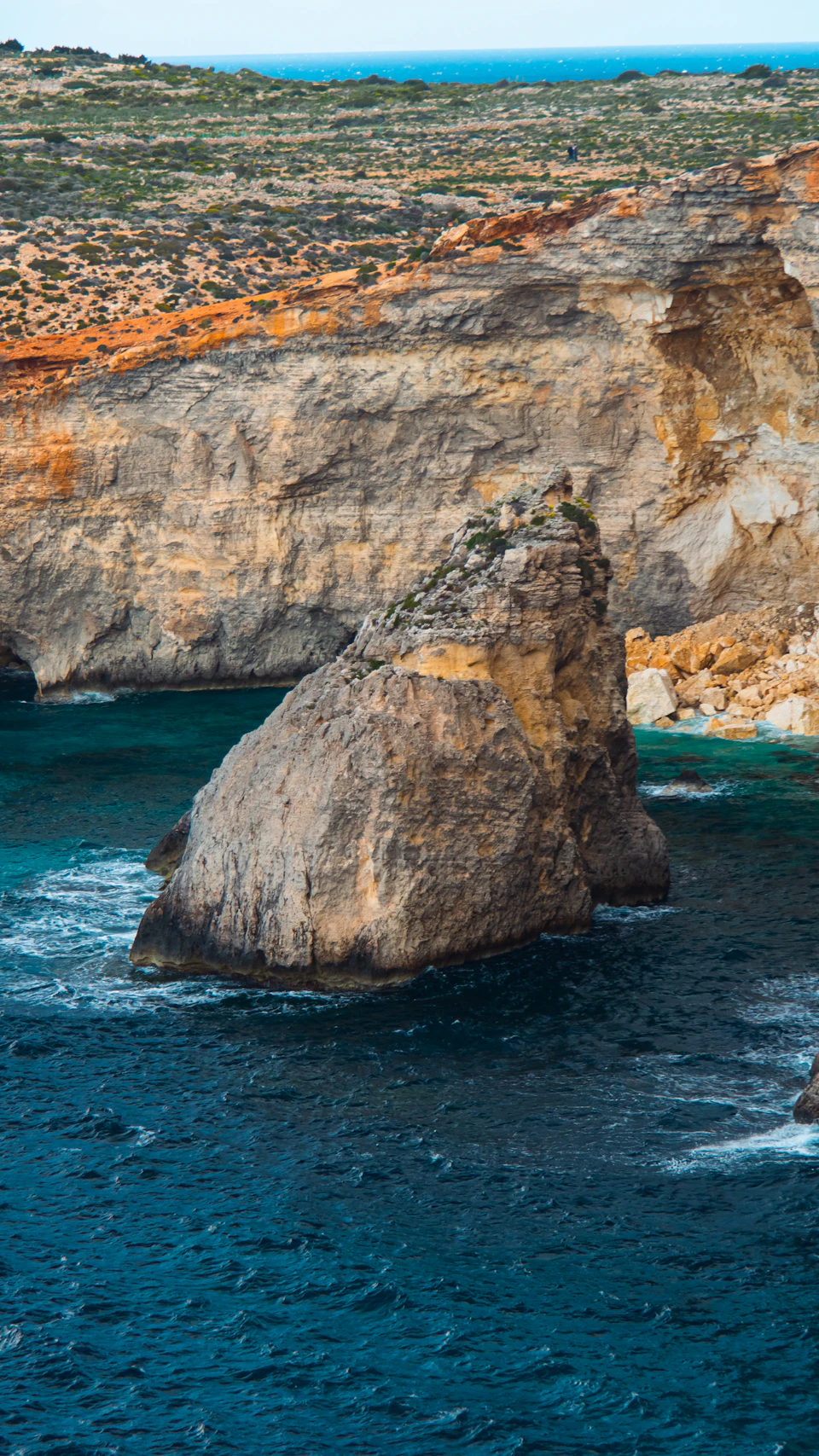 The image depicts a rocky cliff face with a large, jagged rock formation on the right side and a smaller, smoother rock formation on the left side. The cliff is composed of layers of gray and brown rocks, creating a rugged and natural landscape. The water below mirrors the colors of the cliff, with shades of blue and green visible in the foreground.