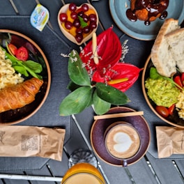 A table setting featuring various breakfast items, including a croissant, scrambled eggs, cherry tomatoes, spinach, and avocado on a plate. A cappuccino with heart-shaped foam is next to a glass of juice. A bowl of cherries and sliced bread are also visible. In the center, there's a vibrant floral arrangement with red flowers and green leaves.