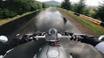 A motorcyclist is riding along a wet road surrounded by lush green trees. The view is from the rider's perspective, showing the motorcycle's handlebars, mirrors, and a smartphone mounted at the center. The sky is overcast, reflecting on the road's surface.