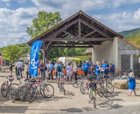 A group of people gathered around a pavilion with bicycles parked nearby. Some individuals are wearing blue shirts, possibly indicating participation in an organized event. A blue flag with writing is visible, and trees and a partially cloudy sky can be seen in the background.