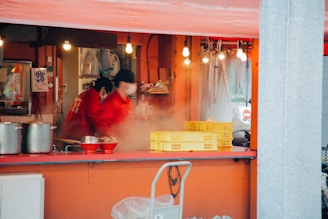 A street food stall with two workers dressed in red jackets and hats. They are busy preparing food, surrounded by steam and various cooking utensils. Yellow plastic crates and metal pots are visible on the counter. The stall is brightly lit with hanging lightbulbs, adding warmth to the atmosphere.