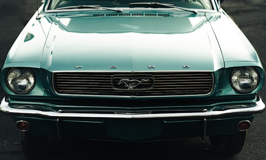 A front view of a vintage Ford Mustang with a teal color, featuring a distinctive grille and round headlights. The classic Mustang logo is prominently displayed at the center of the grille.