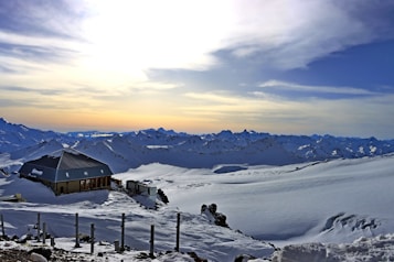 A snowy mountain landscape at sunrise or sunset, featuring a modern cabin with a sloped roof situated on the left side. The sky is a gradient of light orange and blue, enhancing the tranquil atmosphere. Snow-covered peaks stretch across the horizon, and a few posts are visible in the foreground.