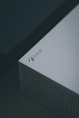 A close-up view of a white gaming console, specifically an Xbox, featuring a perforated design on part of its surface and the Xbox logo clearly visible in the top corner.