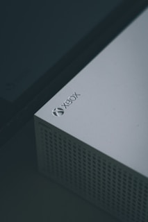 A close-up view of a white gaming console, specifically an Xbox, featuring a perforated design on part of its surface and the Xbox logo clearly visible in the top corner.