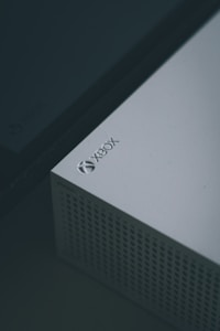 A close-up view of a white gaming console, specifically an Xbox, featuring a perforated design on part of its surface and the Xbox logo clearly visible in the top corner.