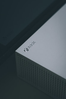 A close-up view of a white gaming console, specifically an Xbox, featuring a perforated design on part of its surface and the Xbox logo clearly visible in the top corner.