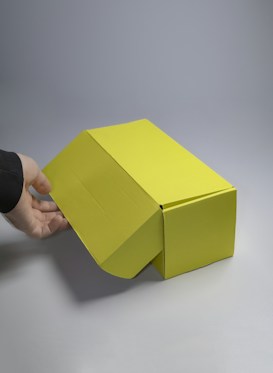 A brightly lit yellow cardboard box with a flap open, revealing a hand reaching towards it. The background is a smooth, neutral gray surface, creating a minimalistic and clean appearance.