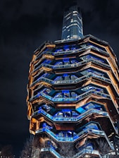 A modern architectural structure with a honeycomb-like pattern is illuminated at night. The design features a series of interconnecting staircases and landings, with blue and orange lighting highlighting its geometric facets. Trees are visible at the base of the structure, and a tall building stands in the background under a dark sky.