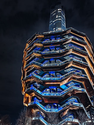 A modern architectural structure with a honeycomb-like pattern is illuminated at night. The design features a series of interconnecting staircases and landings, with blue and orange lighting highlighting its geometric facets. Trees are visible at the base of the structure, and a tall building stands in the background under a dark sky.