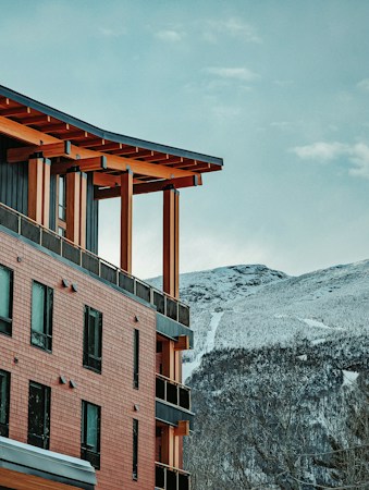A modern building with wooden architectural elements is situated against a backdrop of snow-covered mountains. The structure features brick-like siding, multiple windows, and a slanted roof supported by wooden beams. The sky is clear with a few clouds, giving a serene and natural feeling to the scenery.