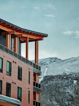 A modern building with wooden architectural elements is situated against a backdrop of snow-covered mountains. The structure features brick-like siding, multiple windows, and a slanted roof supported by wooden beams. The sky is clear with a few clouds, giving a serene and natural feeling to the scenery.