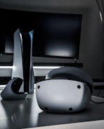 A sleek virtual reality headset sits on a reflective surface beside a gaming console and its controller. The background features a large dark monitor screen, creating a modern and futuristic setting. The lighting is subdued, highlighting the glossy surfaces of the electronic devices.
