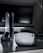 A sleek virtual reality headset sits on a reflective surface beside a gaming console and its controller. The background features a large dark monitor screen, creating a modern and futuristic setting. The lighting is subdued, highlighting the glossy surfaces of the electronic devices.