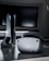 A sleek virtual reality headset sits on a reflective surface beside a gaming console and its controller. The background features a large dark monitor screen, creating a modern and futuristic setting. The lighting is subdued, highlighting the glossy surfaces of the electronic devices.