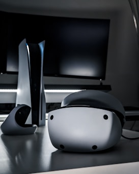 A sleek virtual reality headset sits on a reflective surface beside a gaming console and its controller. The background features a large dark monitor screen, creating a modern and futuristic setting. The lighting is subdued, highlighting the glossy surfaces of the electronic devices.
