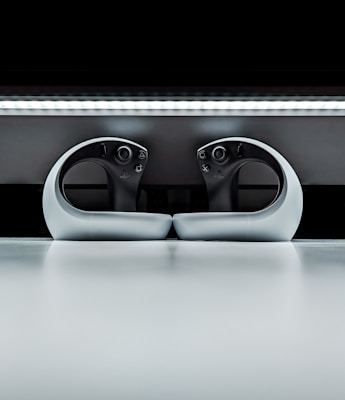 Two sleek, modern gaming controllers with an ergonomic design, placed symmetrically on a reflective surface. The background features a sleek horizontal light strip, enhancing the futuristic appearance.