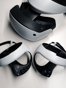 A virtual reality headset and two controllers are neatly arranged on a surface. The headset is predominantly black and white, featuring a sleek design with buttons on the side. The controllers are streamlined with curved grips and feature various buttons on the top side. They are positioned in front of the headset, highlighting their ergonomic shape.