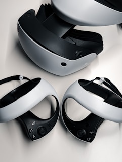 A virtual reality headset and two controllers are neatly arranged on a surface. The headset is predominantly black and white, featuring a sleek design with buttons on the side. The controllers are streamlined with curved grips and feature various buttons on the top side. They are positioned in front of the headset, highlighting their ergonomic shape.