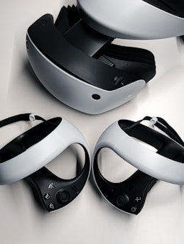 A virtual reality headset and two controllers are neatly arranged on a surface. The headset is predominantly black and white, featuring a sleek design with buttons on the side. The controllers are streamlined with curved grips and feature various buttons on the top side. They are positioned in front of the headset, highlighting their ergonomic shape.