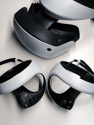 A virtual reality headset and two controllers are neatly arranged on a surface. The headset is predominantly black and white, featuring a sleek design with buttons on the side. The controllers are streamlined with curved grips and feature various buttons on the top side. They are positioned in front of the headset, highlighting their ergonomic shape.