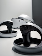 A collection of sleek, modern virtual reality controllers with a circular design are arranged on a flat surface. The controllers have a predominantly black and gray color scheme, featuring buttons and a joystick. The lighting is minimal, creating a futuristic and stylish atmosphere.