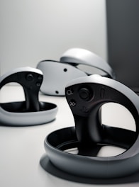 A collection of sleek, modern virtual reality controllers with a circular design are arranged on a flat surface. The controllers have a predominantly black and gray color scheme, featuring buttons and a joystick. The lighting is minimal, creating a futuristic and stylish atmosphere.