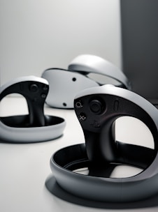 A collection of sleek, modern virtual reality controllers with a circular design are arranged on a flat surface. The controllers have a predominantly black and gray color scheme, featuring buttons and a joystick. The lighting is minimal, creating a futuristic and stylish atmosphere.