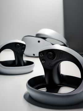 A collection of sleek, modern virtual reality controllers with a circular design are arranged on a flat surface. The controllers have a predominantly black and gray color scheme, featuring buttons and a joystick. The lighting is minimal, creating a futuristic and stylish atmosphere.