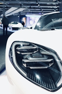 Close-up view of a car's headlight, showcasing a sleek and modern design with precise detailing. The environment appears to be an exhibition or showroom with other cars visible in the blurred background.