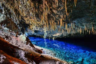 A stunning cave with stalactites hanging from the ceiling and a crystal-clear blue lake reflecting the rock formations above. The cave walls display a variety of textures and colors, with a rugged pathway leading into the depths, partially illuminated by natural light.