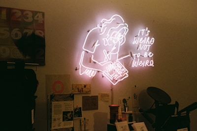 A neon sign on a wall features an outline of a person writing, accompanied by the text 'It's weird not to be weird.' Below the sign, a cluttered workspace includes various papers, notes, and a calendar with numbers. The room has a warm, creative ambiance.