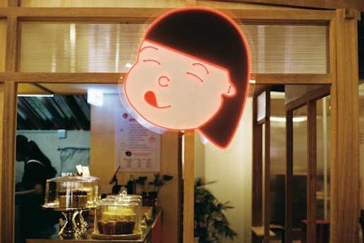 A warm and cozy cafe interior featuring a glowing neon sign of a cartoon girl's face with a cheerful expression. Below the sign, several cake stands display pastries. The wooden decor adds a rustic charm to the space.