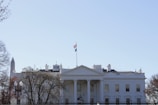A large, neoclassical white building with a prominent facade and a flagpole flying the American flag. The structure is surrounded by trees and a black metal fence, with a clear blue sky above. In the background, a tall, pointed monument is partially visible.