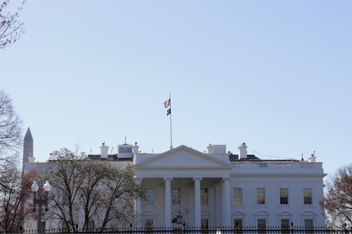 A large, neoclassical white building with a prominent facade and a flagpole flying the American flag. The structure is surrounded by trees and a black metal fence, with a clear blue sky above. In the background, a tall, pointed monument is partially visible.
