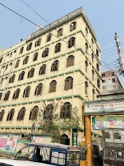 A multi-story building with numerous windows and a sign on top written in a foreign script. Electric wires crisscross in front of the structure. The base of the building features some greenery and there is a blue auto-rickshaw parked nearby. The sky is clear with no visible clouds.