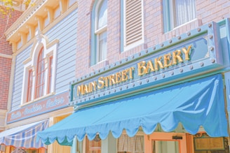 A charming bakery storefront with a brick and pastel blue facade. The lettering on the sign reads 'Main Street Bakery', adorned with small decorative bulbs. The awning is a bright blue, complementing the calm and inviting exterior. Next door, another shop offers fresh goods like bread, cookies, and cakes, as indicated by the signage.