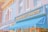 A charming bakery storefront with a brick and pastel blue facade. The lettering on the sign reads 'Main Street Bakery', adorned with small decorative bulbs. The awning is a bright blue, complementing the calm and inviting exterior. Next door, another shop offers fresh goods like bread, cookies, and cakes, as indicated by the signage.