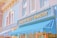 A charming bakery storefront with a brick and pastel blue facade. The lettering on the sign reads 'Main Street Bakery', adorned with small decorative bulbs. The awning is a bright blue, complementing the calm and inviting exterior. Next door, another shop offers fresh goods like bread, cookies, and cakes, as indicated by the signage.
