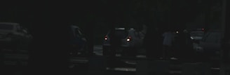Several vehicles are of parked on the side of the road at the evening. A few people are standing near the cars and there is limited visibility due to darkness or low light conditions.