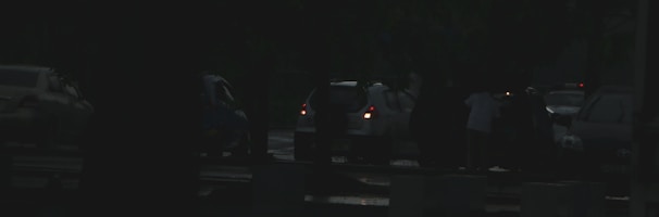Several vehicles are of parked on the side of the road at the evening. A few people are standing near the cars and there is limited visibility due to darkness or low light conditions.