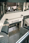 A multi-level market building with a sign reading 'Nam Long Shan Road Cooked Food Market'. The architecture features covered awnings and staircases leading downwards. The interior appears utilitarian with metal railings and tiled flooring.